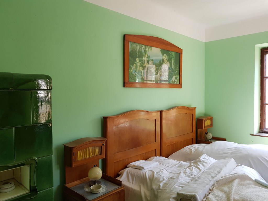 Grünes Gästezimmer im Gasthaus zur Hinterbrühl mit historischen Betten, Nachtkästchen und grünem Kachelofen.